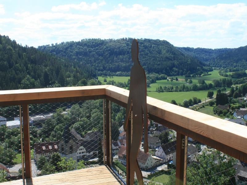 Von einem hölzernen Aussichtspunkt mit Geländer und einer humanoiden Metallskulptur blickt man auf eine grüne, bewaldete Tallandschaft mit einigen Häusern unter einem blauen Himmel mit Wolken.