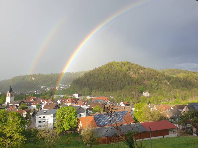 In einem Tal mit einer kleinen Stadt und einer Kirche im Vordergrund spannt sich ein doppelter Regenbogen über einen bewaldeten Hügel.