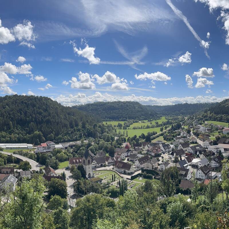 Eine weitläufige Landschaftspanoramaaufnahme zeigt Epfendorf, umgeben von grünen Hügeln und Wäldern unter einem blauen Himmel mit weißen Wolken.