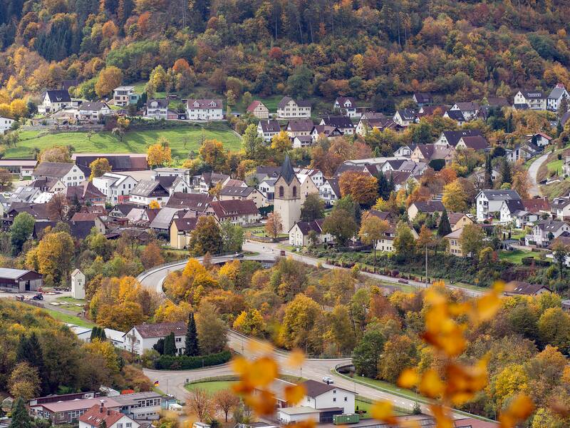 Ein malerisches Dorf mit vielen Häusern und einer Kirche liegt eingebettet in einer herbstlichen, bunt gefärbten Waldlandschaft.