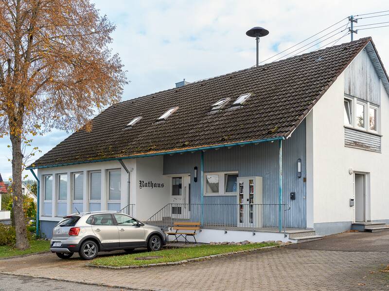 Ein weißes Gebäude mit Schrägdach und mehreren Fenstern hat die Aufschrift "Rathaus" und steht neben einem geparkten Auto und einem Baum.