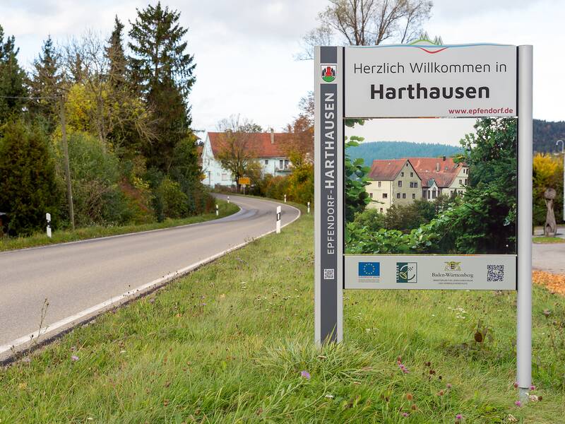 Ein Straßenschild am Rand einer Landstraße begrüßt die Besucher von Harthausen, umgeben von Bäumen und grünen Wiesen.