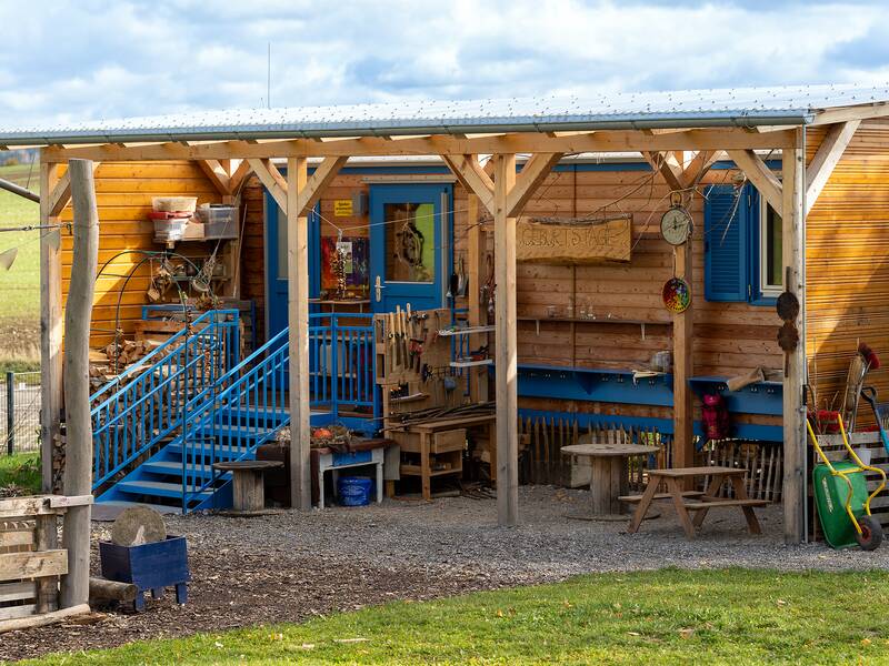 Ein Holzhaus mit blauer Treppe und Überdachung, umgeben von verschiedenem Gartengerät und einem Holztisch mit Bänken.