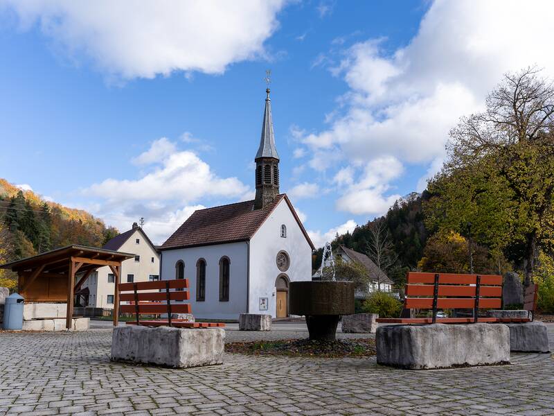 Vor einer kleinen Kirche mit spitzem Turm stehen mehrere Bänke und ein Brunnen auf einem gepflasterten Platz, umgeben von herbstlichen Bäumen und Gebäuden.