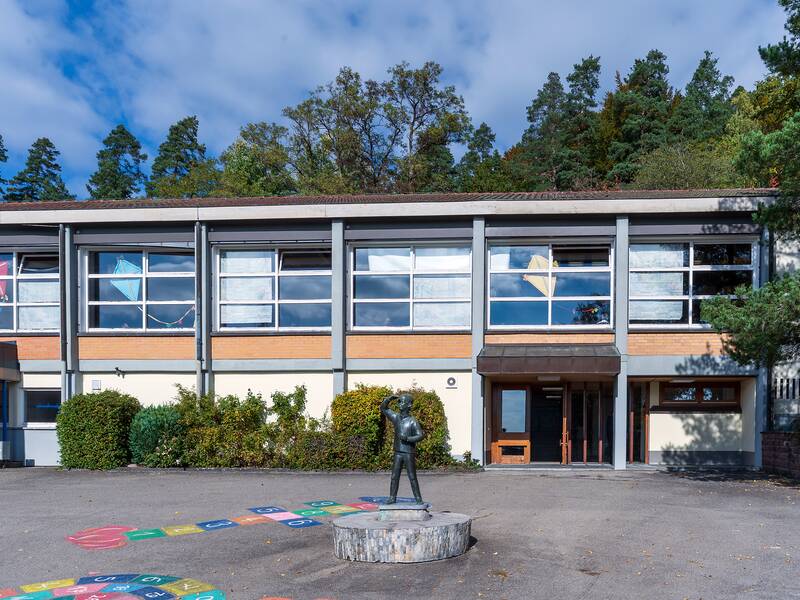 Das Bild zeigt ein zweistöckiges Gebäude mit großen Fenstern und bunten Drachenbildern, umgeben von grünen Bäumen, und einem Spielplatz mit einer Statue im Vordergrund.