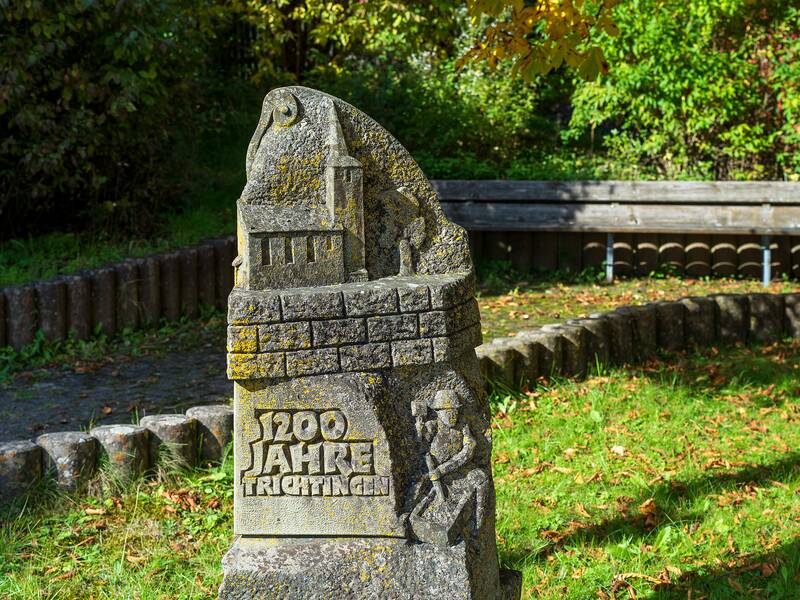 Ein verwitterter Steinsockel mit der Inschrift "1200 Jahre Trichtingen" und einem eingravierten Gebäudemotiv steht auf einer Rasenfläche, umgeben von Bäumen und einer Holzbank.