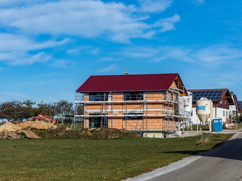 Neu gebautes Haus, das noch eingerüstet ist. Das Haus steht am Ende eines Wohngebiets auf einer grünen Wiese. Der Himmel ist blau.